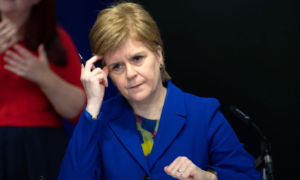 Scotland's first minister Nicola Sturgeon wears a blue and green top with a matching blue jacket during a press conference. Her head is resting against her hand