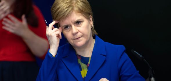 Scotland's first minister Nicola Sturgeon wears a blue and green top with a matching blue jacket during a press conference. Her head is resting against her hand
