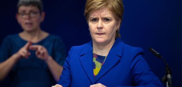 Scotland's first minister Nicola Sturgeon wears a blue and green top with a matching blue jacket during a press conference. She is standing at a podium