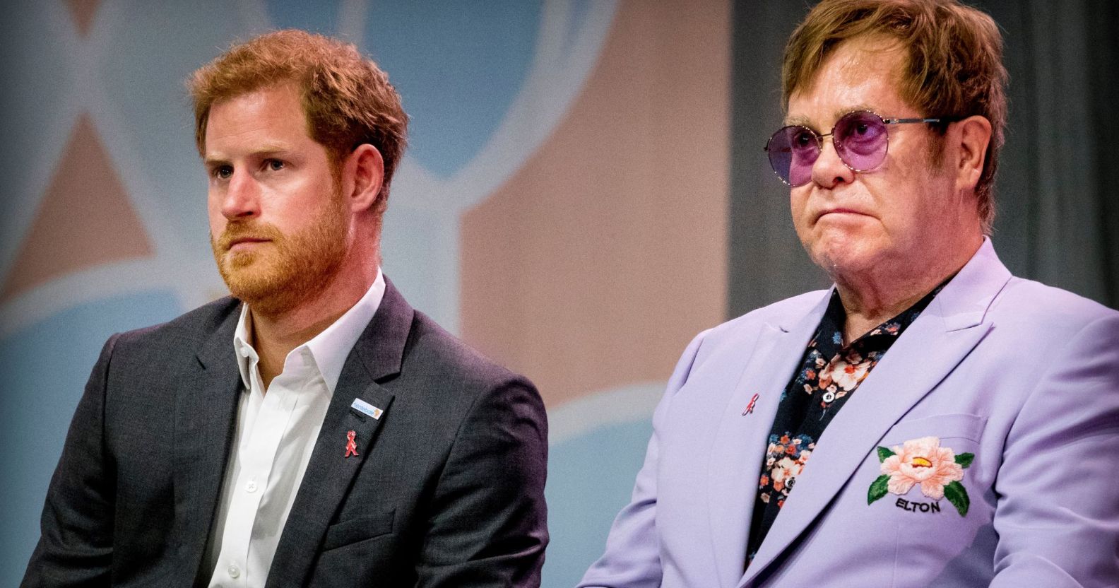Prince Harry wears a white shirt and dark grey suit jacket as he sits next to Elton John, who is wearing a light purple jacket and dark shirt with floral patterning on it