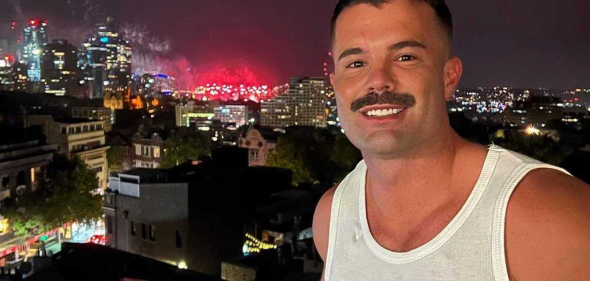 Simon, a white man with a moustache, smiling while stood on a balcony in front of a nighttime skyline