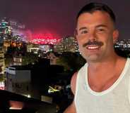Simon, a white man with a moustache, smiling while stood on a balcony in front of a nighttime skyline