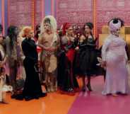 A screenshot from RuPaul’s Drag Race season 15 shows the drag queen contestants standing next to each other in one of the series' sets that has a orange and pink floor with pink-pattern walls and lights in the background