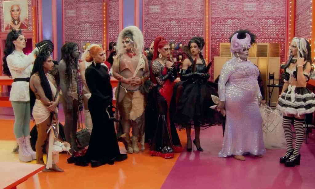 A screenshot from RuPaul’s Drag Race season 15 shows the drag queen contestants standing next to each other in one of the series' sets that has a orange and pink floor with pink-pattern walls and lights in the background