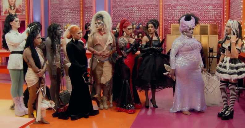 A screenshot from RuPaul’s Drag Race season 15 shows the drag queen contestants standing next to each other in one of the series' sets that has a orange and pink floor with pink-pattern walls and lights in the background