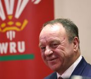Steve Phillips wears a white button-up shirt and blue jacket as he stands in front of a red banner displaying the Welsh Rugby Union logo and WRU lettering