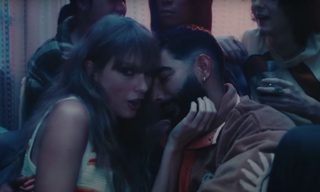 Taylor Swift looks towards the camera as she holds the face of Laith Ashley, a trans model and singer, in a house party scene in her "Lavender Haze" music video