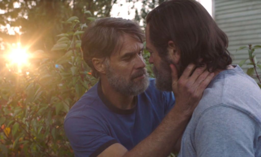 screenshot of Bill and Frank in The Last of Us shows their characters played by Nick Offerman and Murray Bartlett embracing as sunlight can be seen shining through their garden on the left-hand side of the screen