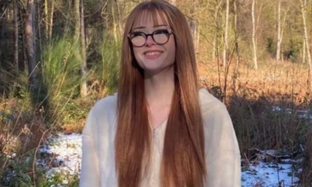 Brianna Ghey pictured outdoors wearing a light-coloured hoodie. Her hair is long and auburn and she is smiling and wearing thick-rimmed glasses. Trees can be seen in the background.