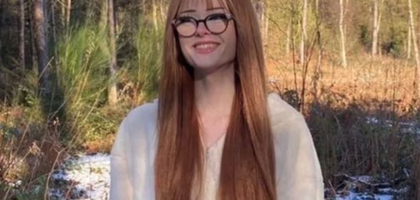 Brianna Ghey pictured outdoors wearing a light-coloured hoodie. Her hair is long and auburn and she is smiling and wearing thick-rimmed glasses. Trees can be seen in the background.
