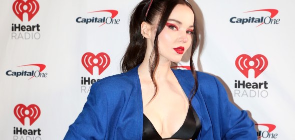 Dove Cameron wearing a blue blazer and black bralet with her hair tied up and red eye makeup on a white background.