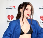 Dove Cameron wearing a blue blazer and black bralet with her hair tied up and red eye makeup on a white background.