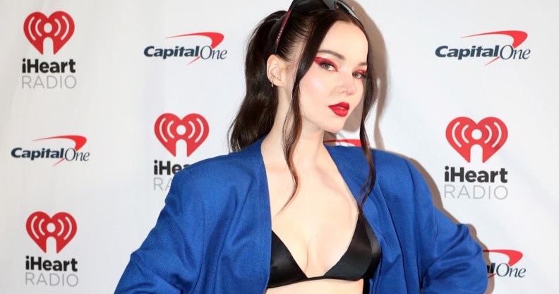 Dove Cameron wearing a blue blazer and black bralet with her hair tied up and red eye makeup on a white background.