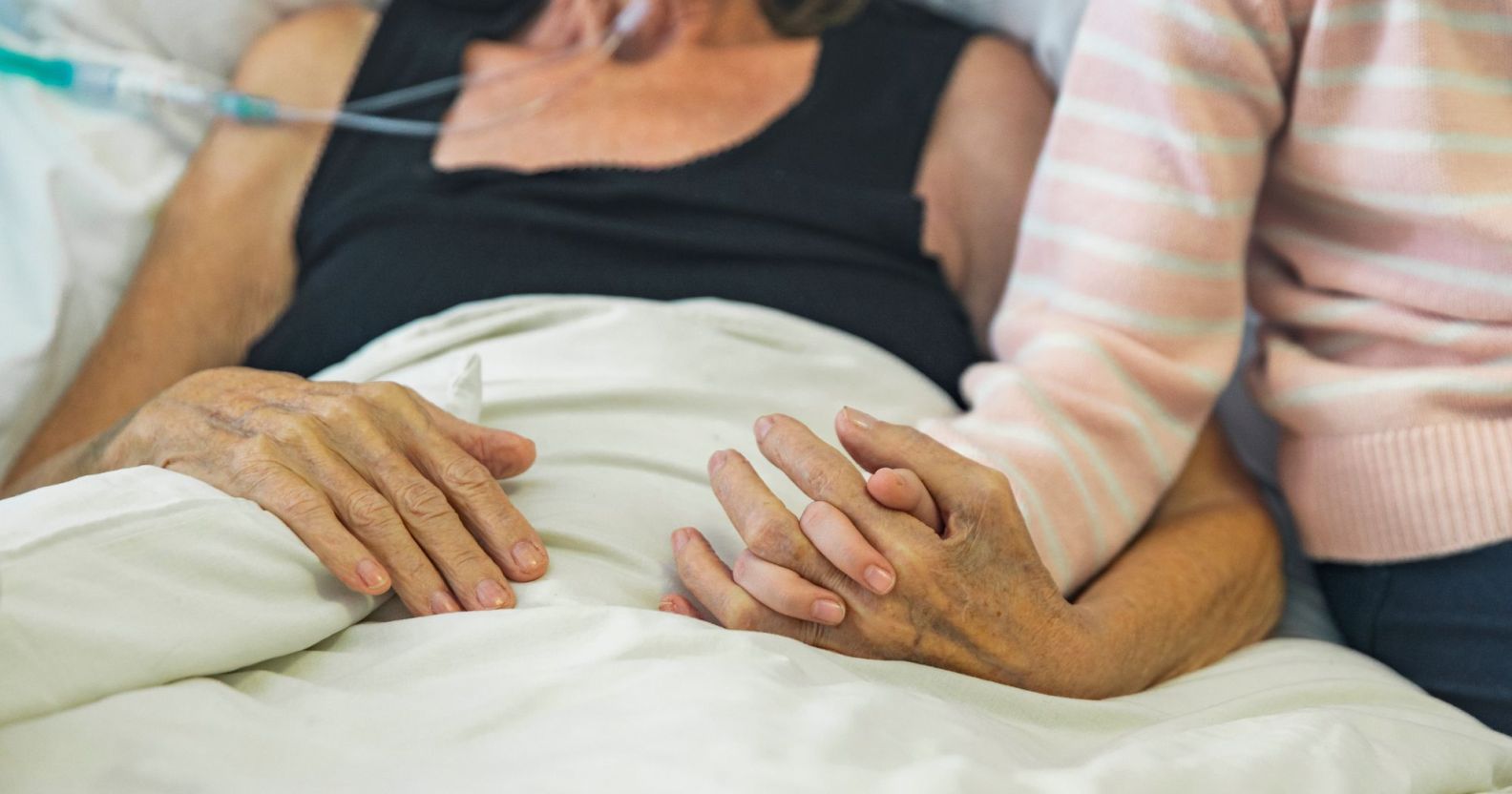 A bed-ridden person holds hands with a loved one.