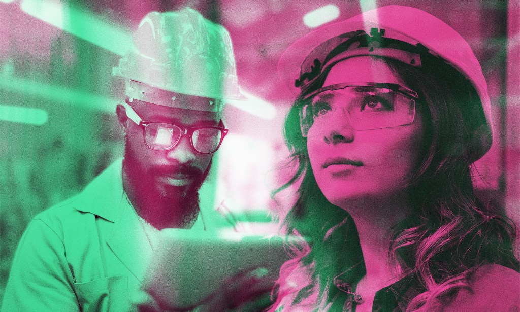 A man in a hard hat look down at a tablet while a woman with safety glasses and a hard hat looks on.