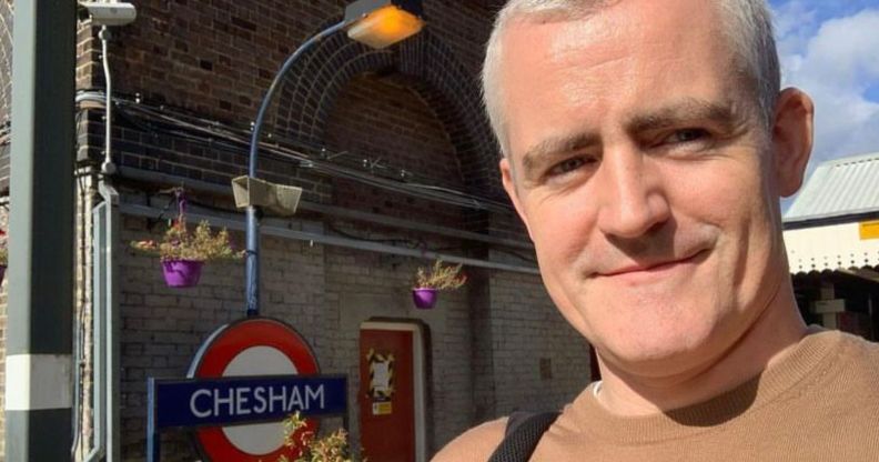Eugene Lynch pictured at an overground station in London. He is on the right hand side of the image and has grey hair and is wearing a brown t-shirt. An underground sign can be seen in the background.