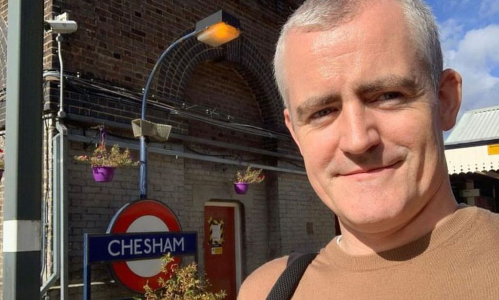 Eugene Lynch pictured at an overground station in London. He is on the right hand side of the image and has grey hair and is wearing a brown t-shirt. An underground sign can be seen in the background.