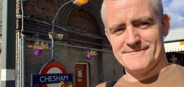 Eugene Lynch pictured at an overground station in London. He is on the right hand side of the image and has grey hair and is wearing a brown t-shirt. An underground sign can be seen in the background.