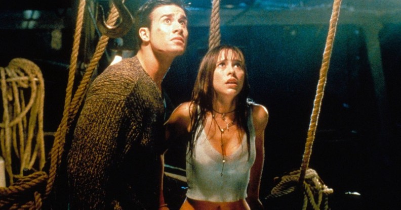 A screenshot from I Know What You Did Last Summer shows actor Freddie Prinze Jr wearing a dark top standing next to actor Jennifer Love Hewitt wearing a white vest top as they both look scared while they stand inside the dark interior of a boat