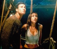 A screenshot from I Know What You Did Last Summer shows actor Freddie Prinze Jr wearing a dark top standing next to actor Jennifer Love Hewitt wearing a white vest top as they both look scared while they stand inside the dark interior of a boat