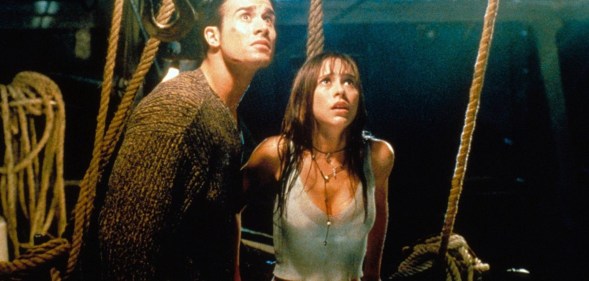 A screenshot from I Know What You Did Last Summer shows actor Freddie Prinze Jr wearing a dark top standing next to actor Jennifer Love Hewitt wearing a white vest top as they both look scared while they stand inside the dark interior of a boat