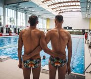 Two men wait for the results of their dive at the 2018 Gay Games. They stand with their arms around each other and they are pictured from behind standing beside a swimming pool.