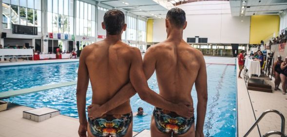 Two men wait for the results of their dive at the 2018 Gay Games. They stand with their arms around each other and they are pictured from behind standing beside a swimming pool.