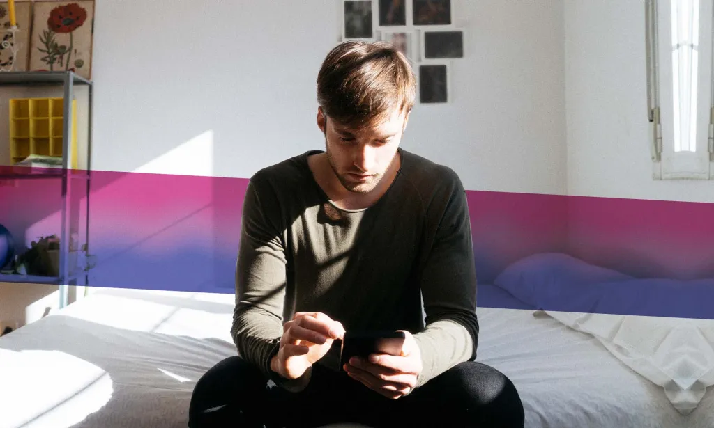 Man sitting on bed looking at phone with bisexual Pride flag colours behind him