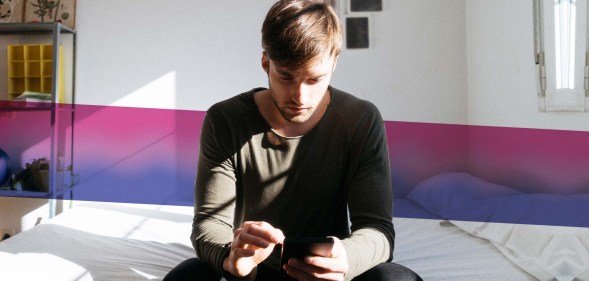 Man sitting on bed looking at phone with bisexual Pride flag colours behind him