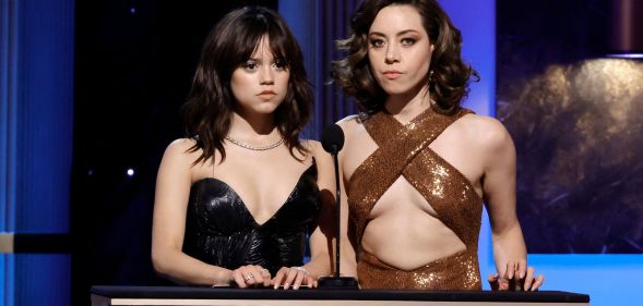 Jenna Ortega in a sparkly black top and Aubrey Plaza in a gold cross strap top, presenting at the 2023 SAG Award. Both actresses have a deadpan expression.