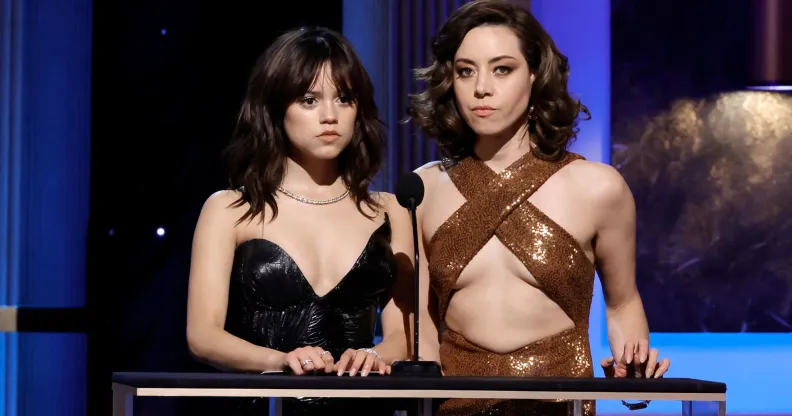 Jenna Ortega in a sparkly black top and Aubrey Plaza in a gold cross strap top, presenting at the 2023 SAG Award. Both actresses have a deadpan expression.