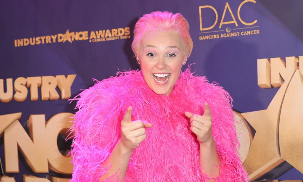 JoJo Siwa on the 2022 Industry Dance Awards red carpet, wearing a pink fluffy jacket and slicked back blonde hair. JoJo is smiling and pointing at the camera.