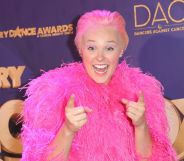 JoJo Siwa on the 2022 Industry Dance Awards red carpet, wearing a pink fluffy jacket and slicked back blonde hair. JoJo is smiling and pointing at the camera.