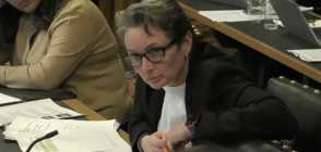 A screenshot of Labour MP Kate Osbourne wearing a white shirt and black suit jacket as she sits at a desk with papers in front of her during a women and equalities committee meeting