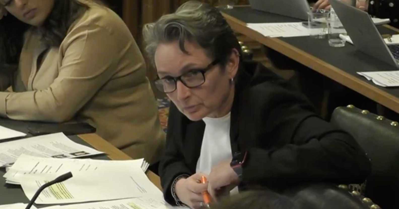 A screenshot of Labour MP Kate Osbourne wearing a white shirt and black suit jacket as she sits at a desk with papers in front of her during a women and equalities committee meeting
