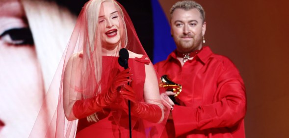 A screenshot of trans artist Kim Petras on stage at the Grammy awards with non-binary singer Sam Smith. The pair are dressed in red outfits, in the background you can see part of a large projected face of Kim Petras (Getty)