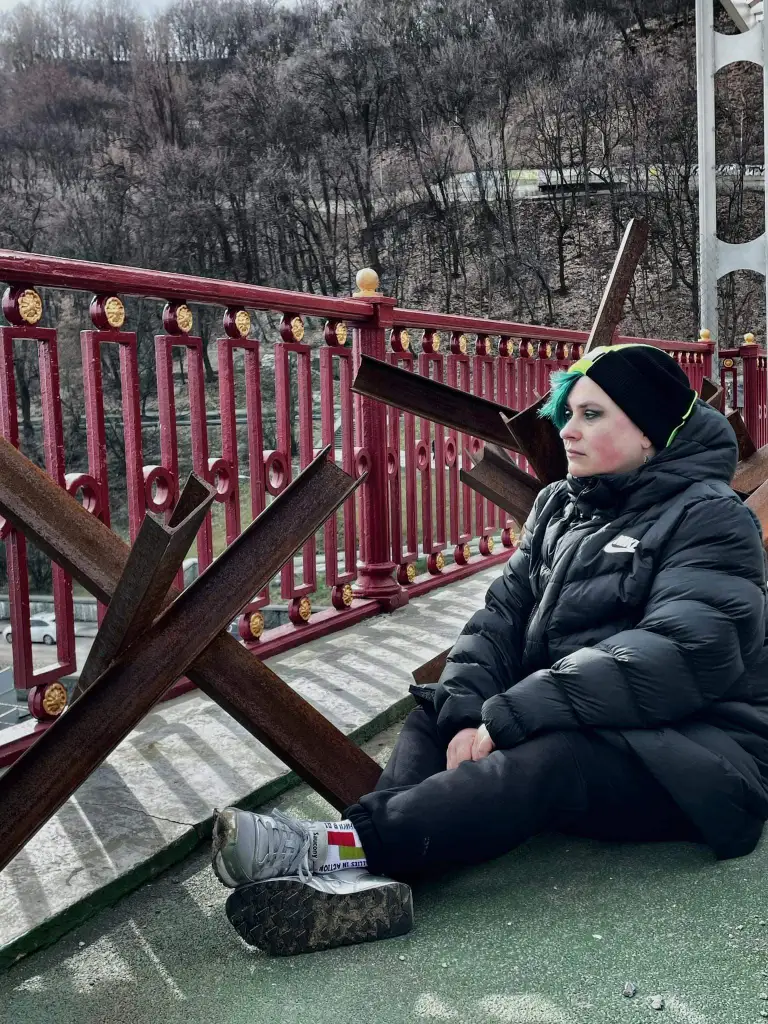 Edward Reese of Kyiv Pride. He is pictured outdoors sitting on the ground by a fence wearing a black puffer jacket. He has green hair.