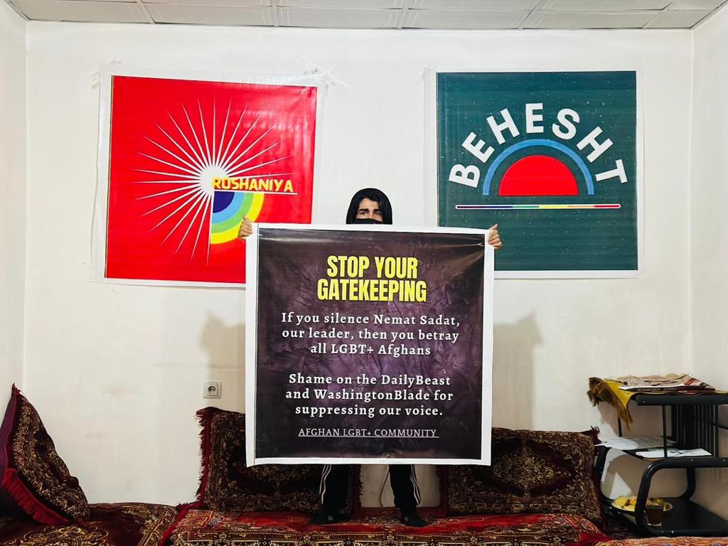 A member of Afghanistan's LGBTQ+ group Behesht Collective stands holding a poster which reads "Stop the gatekeeping". In the background are two posters on a awl advertising Roshaniya and the Behesht Collective.
