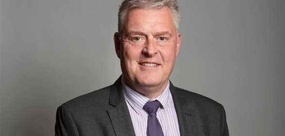 Lee Anderson pictured in his official parliamentary portrait. He is wearing a grey suit with a dark coloured tie and is standing against a grey background.