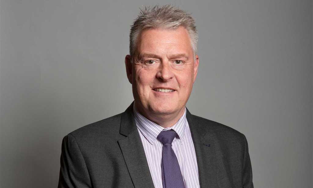 Lee Anderson pictured in his official parliamentary portrait. He is wearing a grey suit with a dark coloured tie and is standing against a grey background.