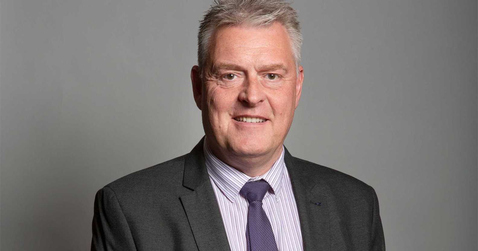 Lee Anderson pictured in his official parliamentary portrait. He is wearing a grey suit with a dark coloured tie and is standing against a grey background.