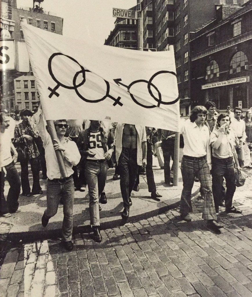 Mark Segal (far-right) at an LGBTQ+ equality march beside a flag with two interlocked male and female symbols