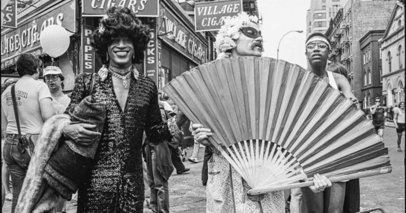 Marsha P Johnson pictured at a march in the early '70s. She is pictured wearing a long dress and a hat besides two activists who are holding up a giant fan. They are pictured on the streets of New York.
