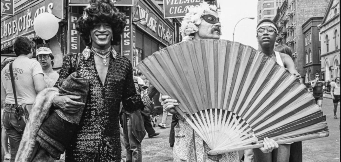 Marsha P Johnson pictured at a march in the early '70s. She is pictured wearing a long dress and a hat besides two activists who are holding up a giant fan. They are pictured on the streets of New York.