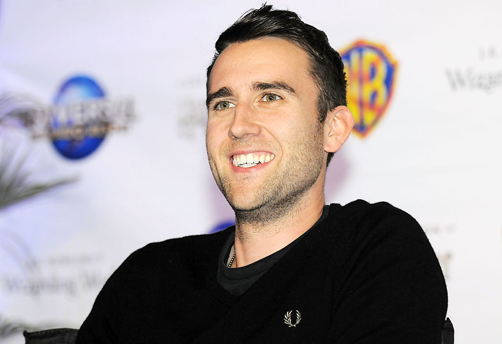 Matthew Lewis smiling in a black jumper in front of a Warner Brothers logo