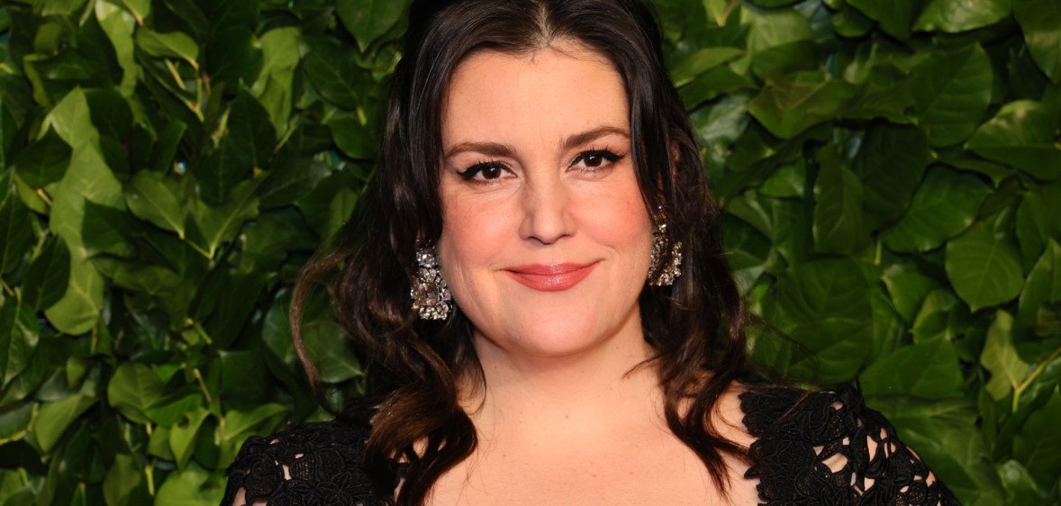 Photo of actor Melanie Lynskey wearing a black dress smiling set against a background of green leaves