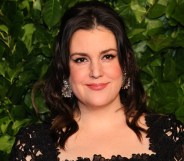 Photo of actor Melanie Lynskey wearing a black dress smiling set against a background of green leaves