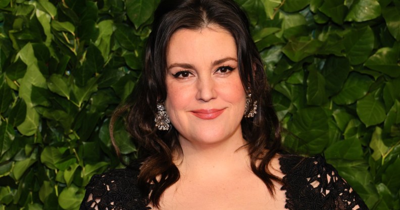 Photo of actor Melanie Lynskey wearing a black dress smiling set against a background of green leaves