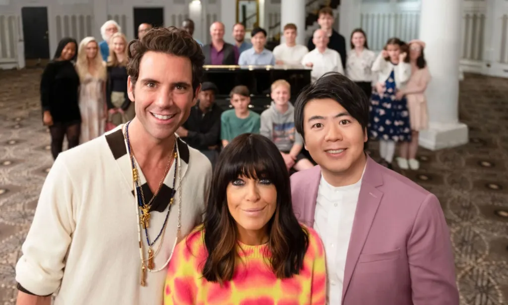 Mika, Claudia Winkleman and pianist Lang Lang stand in the foreground looking at the camera, while a group of piano players stand in the background next to a piano.