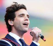 Singer Mika wearing a blue, white and red suit, white shirt and blue tie while performing on stage, with a grey microphone in his hand.